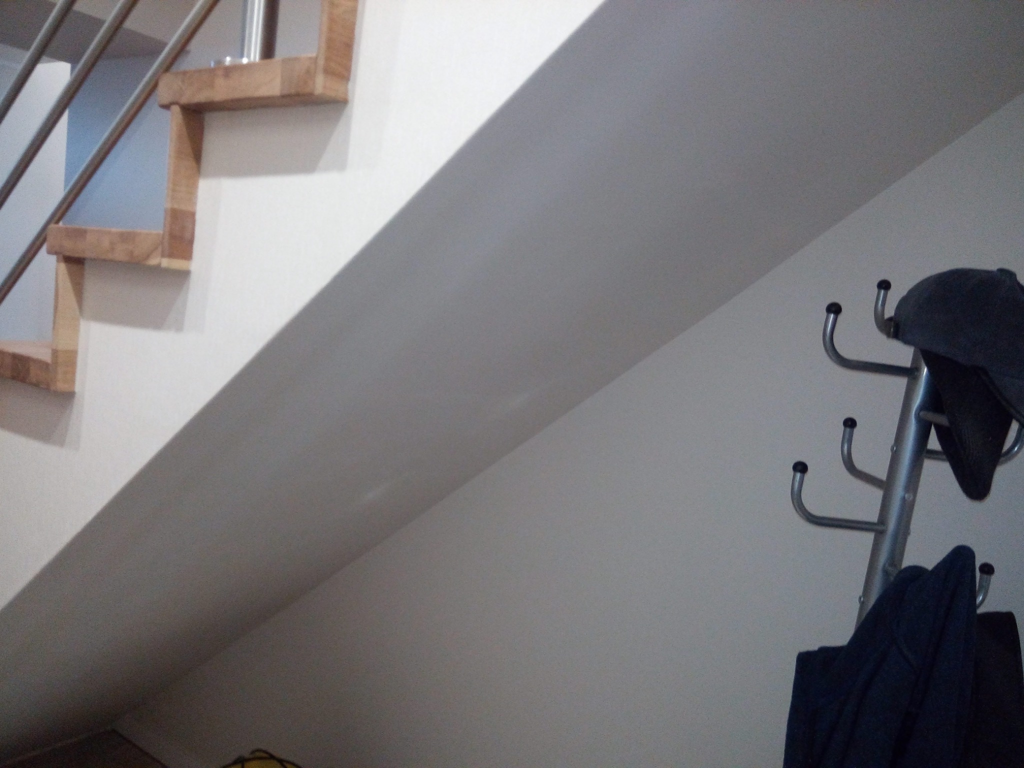 Fragment klatki schodowej z drewnianymi stopniami i metalową balustradą, widok na białą ścianę i wieszak na ubrania z ciemnymi ubraniami.