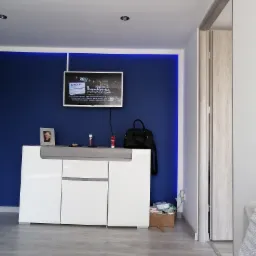 Nowoczesny salon z granatową ścianą akcentującą, podświetloną listwą LED, białą komodą z szarym blatem, telewizorem i drewnianą podłogą, widok na otwarte drzwi do innego pomieszczenia.