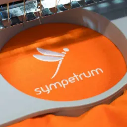 Pomarańczowa tkanina w tamborku z wyhaftowanym białym logo i napisem 'sympetrum', widoczna głowica maszyny do haftowania z igłami.