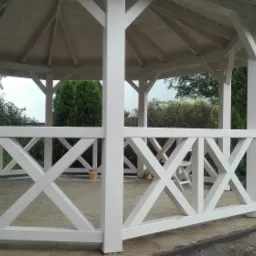 Biała, drewniana altana z balustradą w kształcie krzyży, widok z zewnątrz na zieleń ogrodu i białą przyczepę w tle.