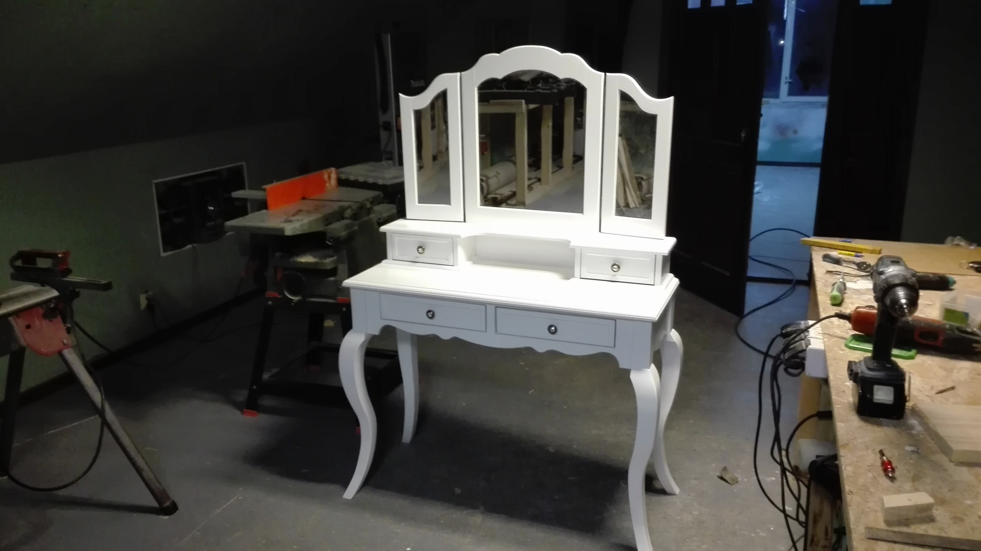 Biała toaletka z trzema lustrami w stylu retro, z szufladami i giętymi nogami, ustawiona w warsztacie stolarskim z widocznymi narzędziami i maszynami.