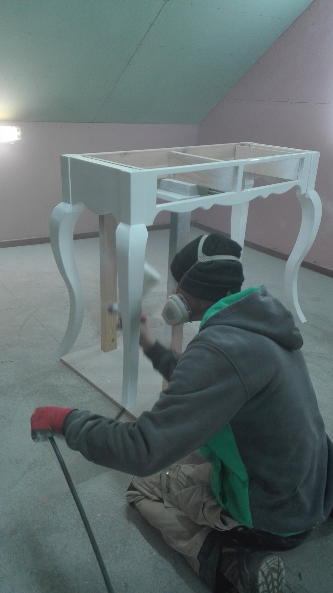 Pracownik w masce i rękawicach maluje natryskowo białą farbą drewniany stół z giętymi nogami w surowym pomieszczeniu z szarym betonem na podłodze i różowymi ścianami.