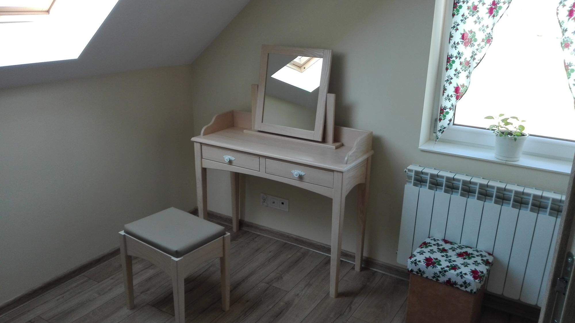 Jasna, drewniana toaletka z lustrem i krzesłem w pokoju na poddaszu, obok okna z kwiecistą zasłoną i grzejnikiem.