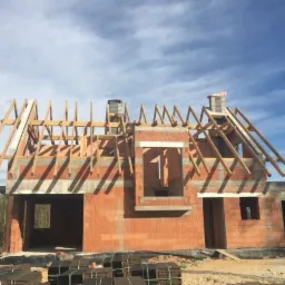 Surowy stan budowy domu z czerwonej cegły z drewnianą konstrukcją dachu w trakcie montażu, widoczne pustaki ceramiczne i elementy więźby dachowej na tle błękitnego nieba.