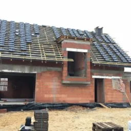 Dom w trakcie budowy z częściowo ułożonymi dachówkami w kolorze grafitowym, widoczne łaty dachowe i komin. Budynek z czerwonej cegły, otwarty stan surowy.