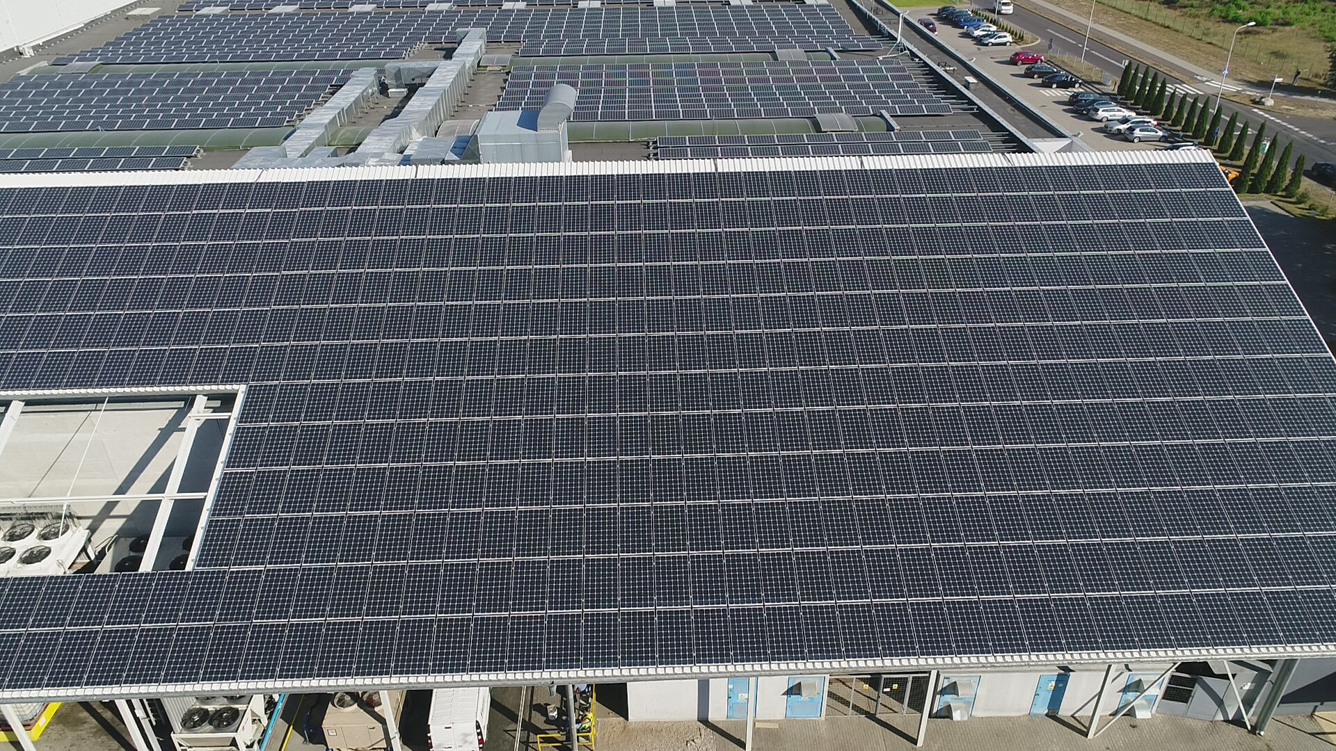Widok z drona na rozległą instalację paneli słonecznych na dachu budynku przemysłowego, z widocznymi jednostkami klimatyzacyjnymi i zaparkowanymi samochodami w tle.