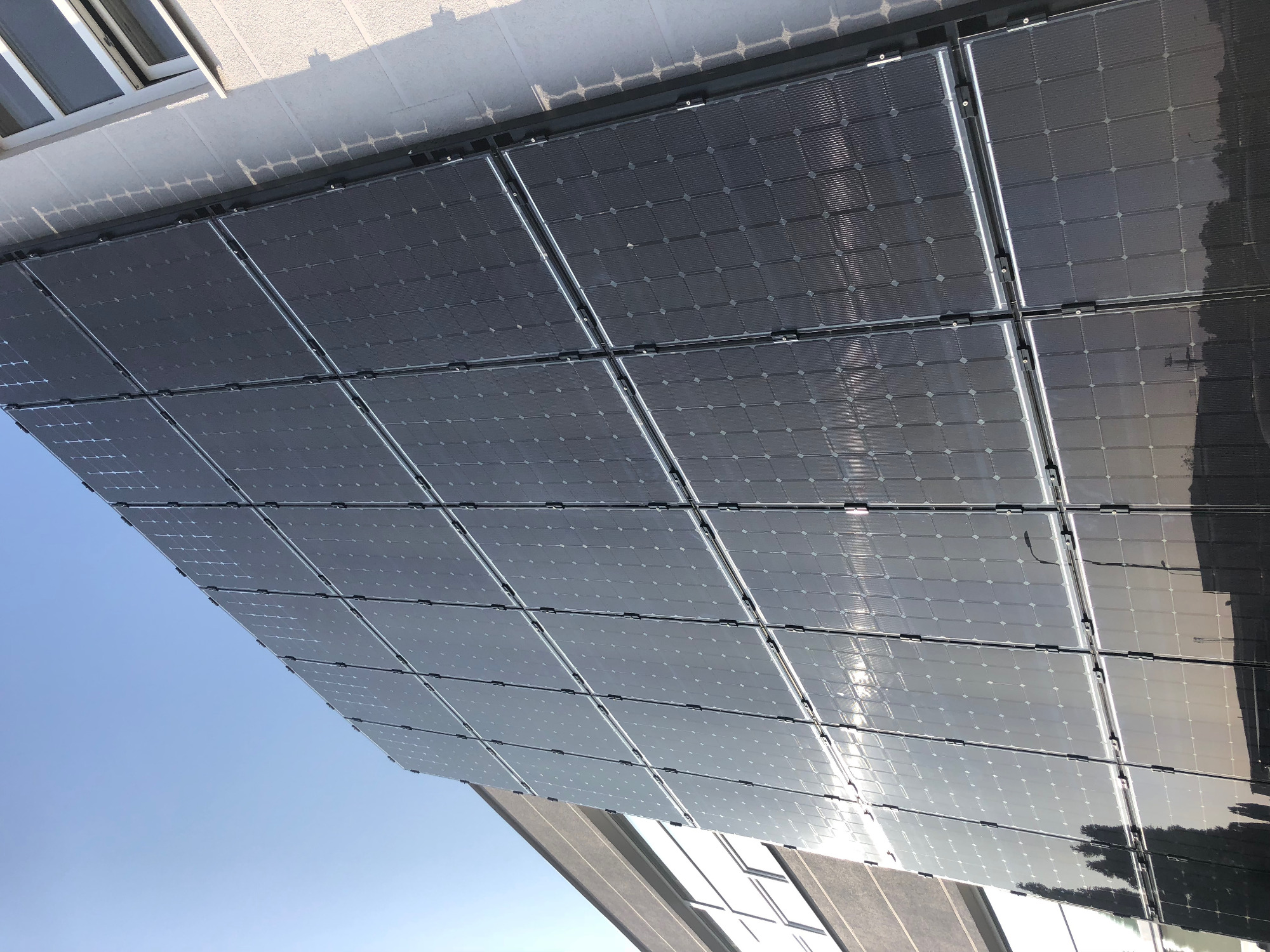 Instalacja fotowoltaiczna na dachu budynku, widok od dołu pod kątem, panele w czarnych ramach odbijają błękitne niebo.