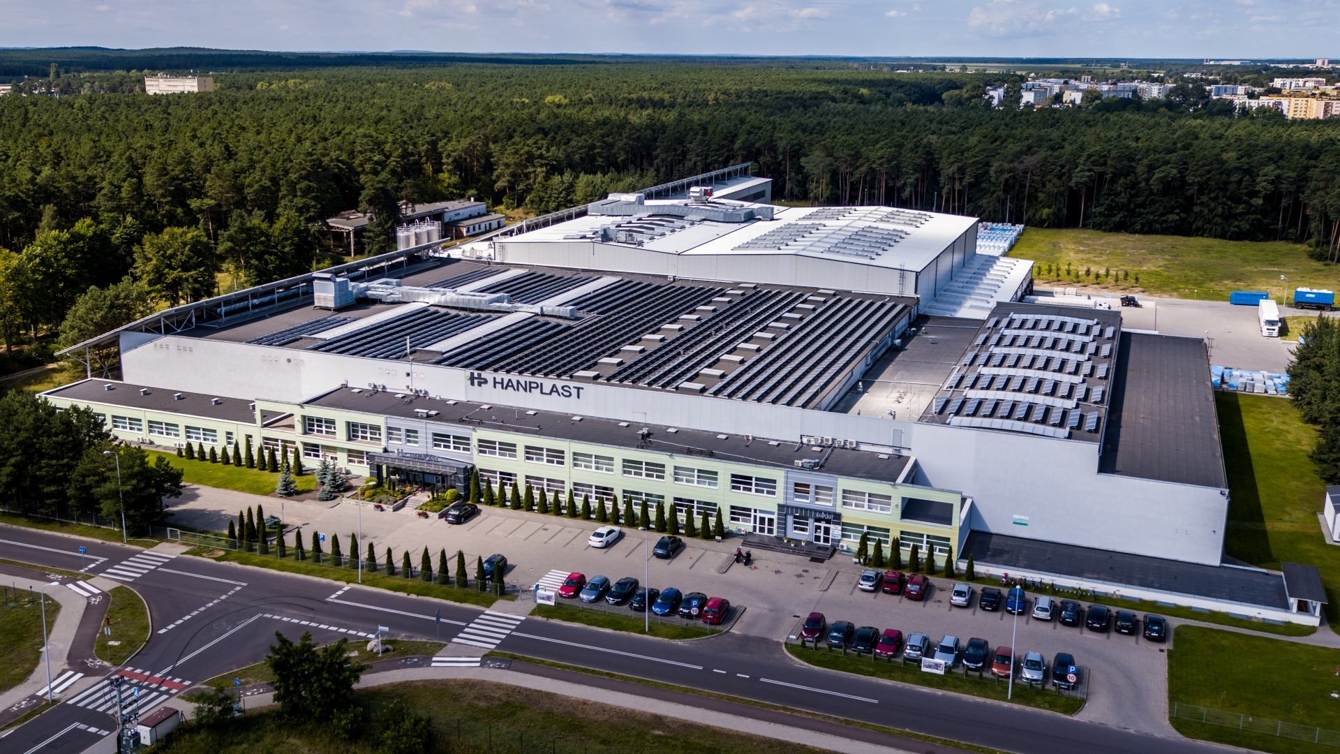 Widok z lotu ptaka na dużą halę produkcyjną Hanplast z zainstalowanymi panelami słonecznymi na dachu, otoczoną zielenią i parkingiem z samochodami.