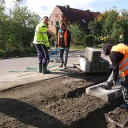 Trzech pracowników w odblaskowych kamizelkach instaluje betonowe krawężniki przy drodze, używając specjalistycznych narzędzi do podnoszenia i ustawiania elementów.