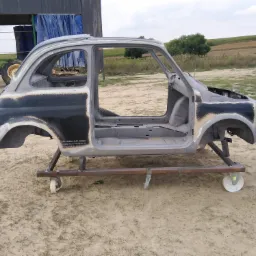 Karoseria Fiata 500 bez drzwi i szyb, osadzona na metalowym wózku na kółkach, przygotowana do renowacji na zewnątrz.