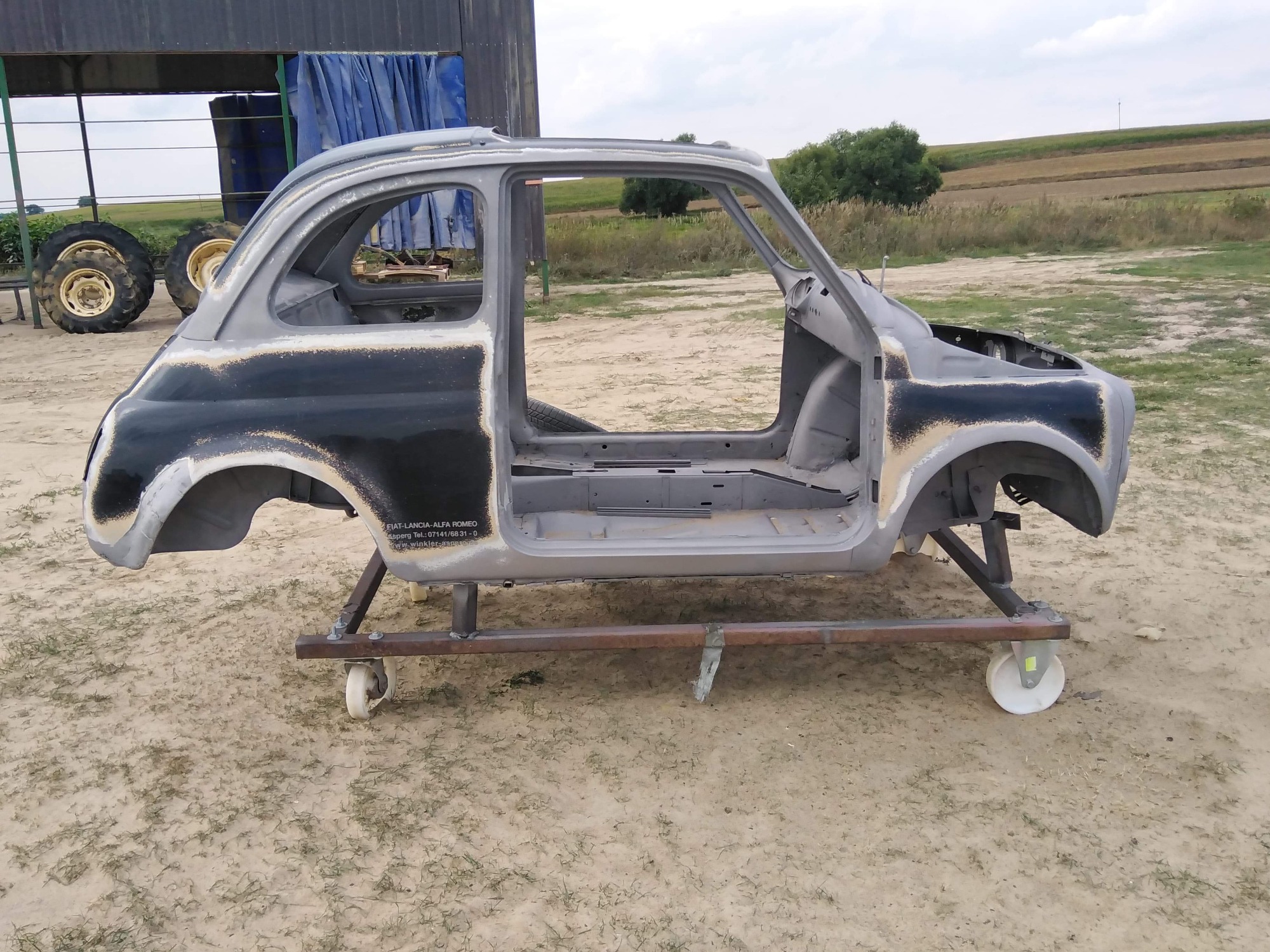 Karoseria Fiata 500 bez drzwi i szyb, osadzona na metalowym wózku na kółkach, przygotowana do renowacji na zewnątrz.