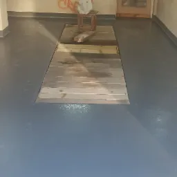 Szara, gładka podłoga w pomieszczeniu z surowymi ścianami, fragment drewnianej rampy i stołek w tle.