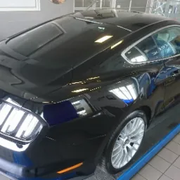 Czarny samochód marki Ford Mustang na podnośniku w warsztacie, widok z tyłu, z widocznymi refleksami światła na karoserii i felgach.