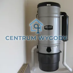 Srebrno-czarny system centralnego odkurzania marki Beam zamontowany na jasnej ścianie, widoczne rury instalacyjne i fragment podłogi z panelami.