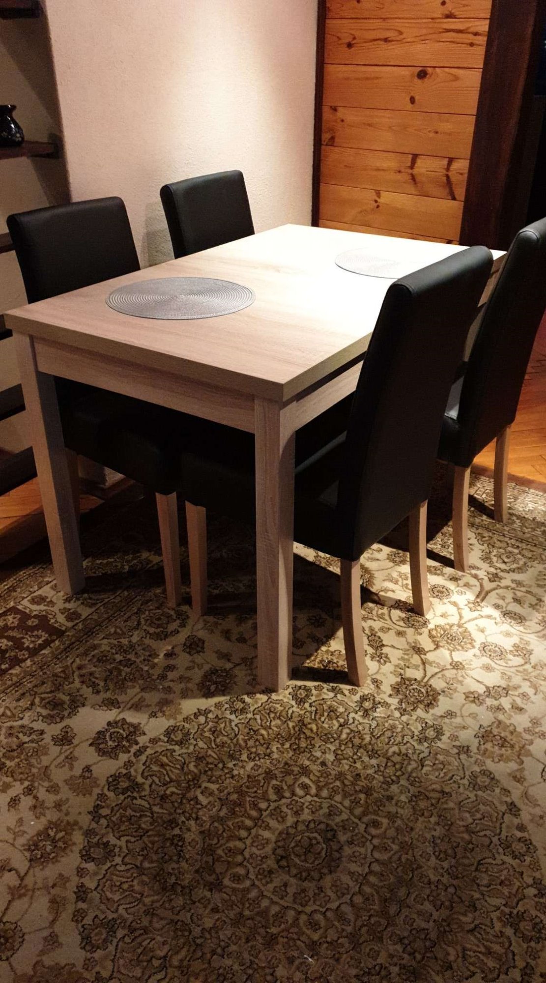 Jasny, drewniany stół z czterema czarnymi krzesłami na wzorzystym dywanie, widok z niskiego kąta.
