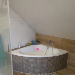 Narożna wanna obudowana mozaiką w łazience na poddaszu, z widoczną kabiną prysznicową, drewnopodobnymi płytkami i dywanikiem geometrycznym.