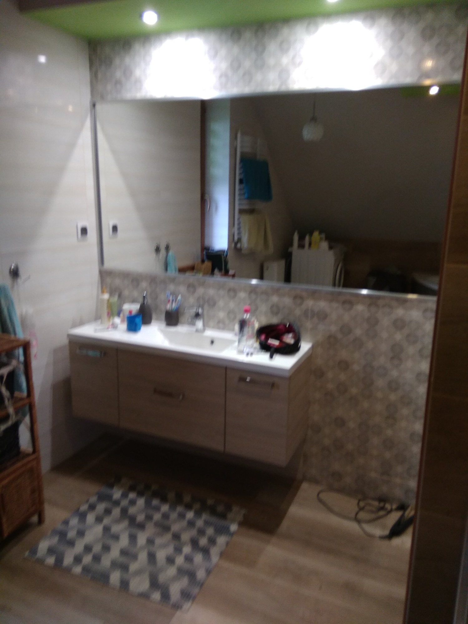 Nowoczesna łazienka z podwieszaną szafką pod umywalką, dużym lustrem i wzorzystą tapetą, widok na odbicie w lustrze ukazujące skos dachu.