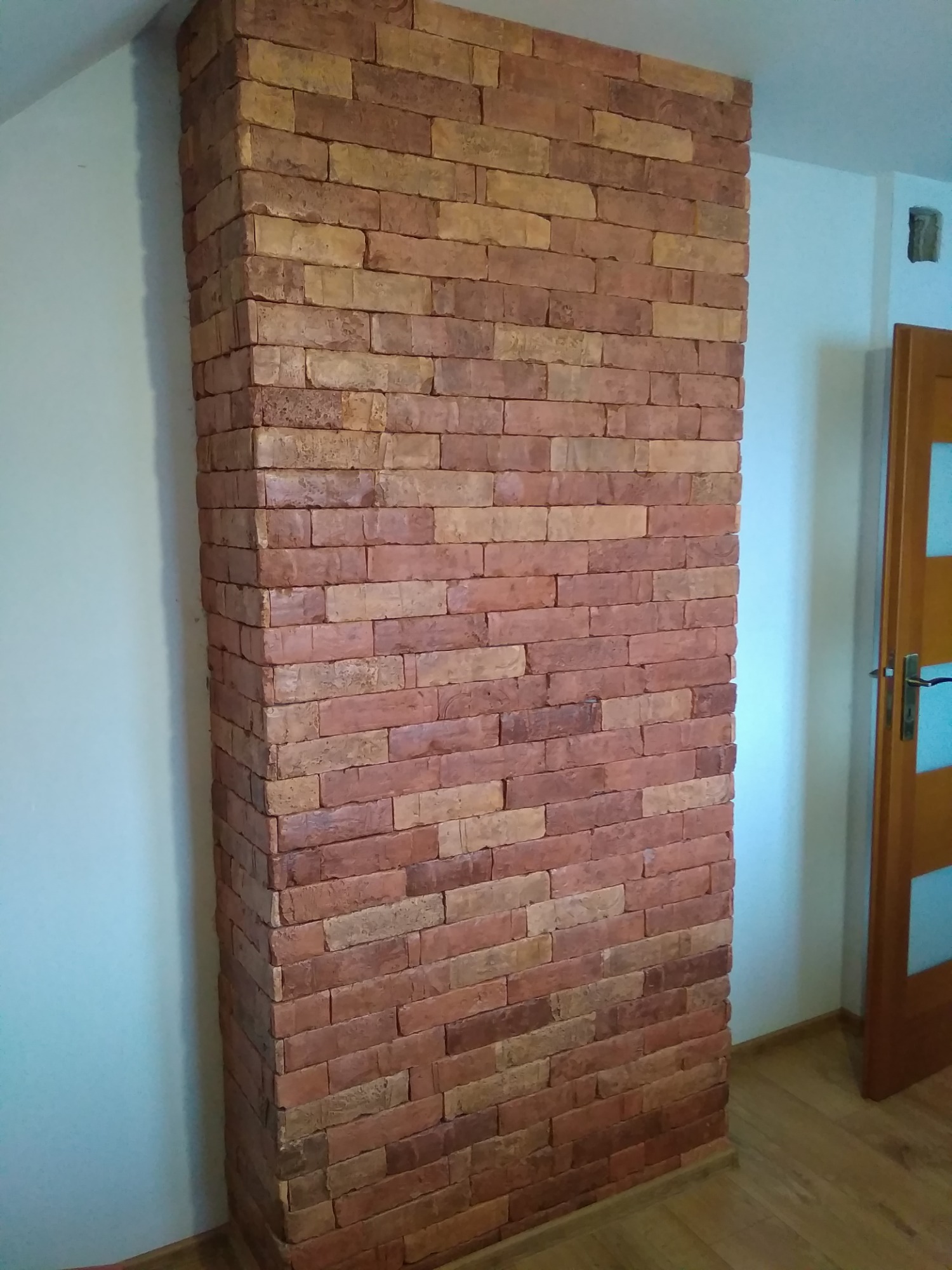 Ściana wewnętrzna wyłożona cegłą dekoracyjną w odcieniach czerwieni i beżu, kontrastująca z gładką, białą ścianą i drewnianą podłogą, widoczny fragment ościeżnicy drzwi.