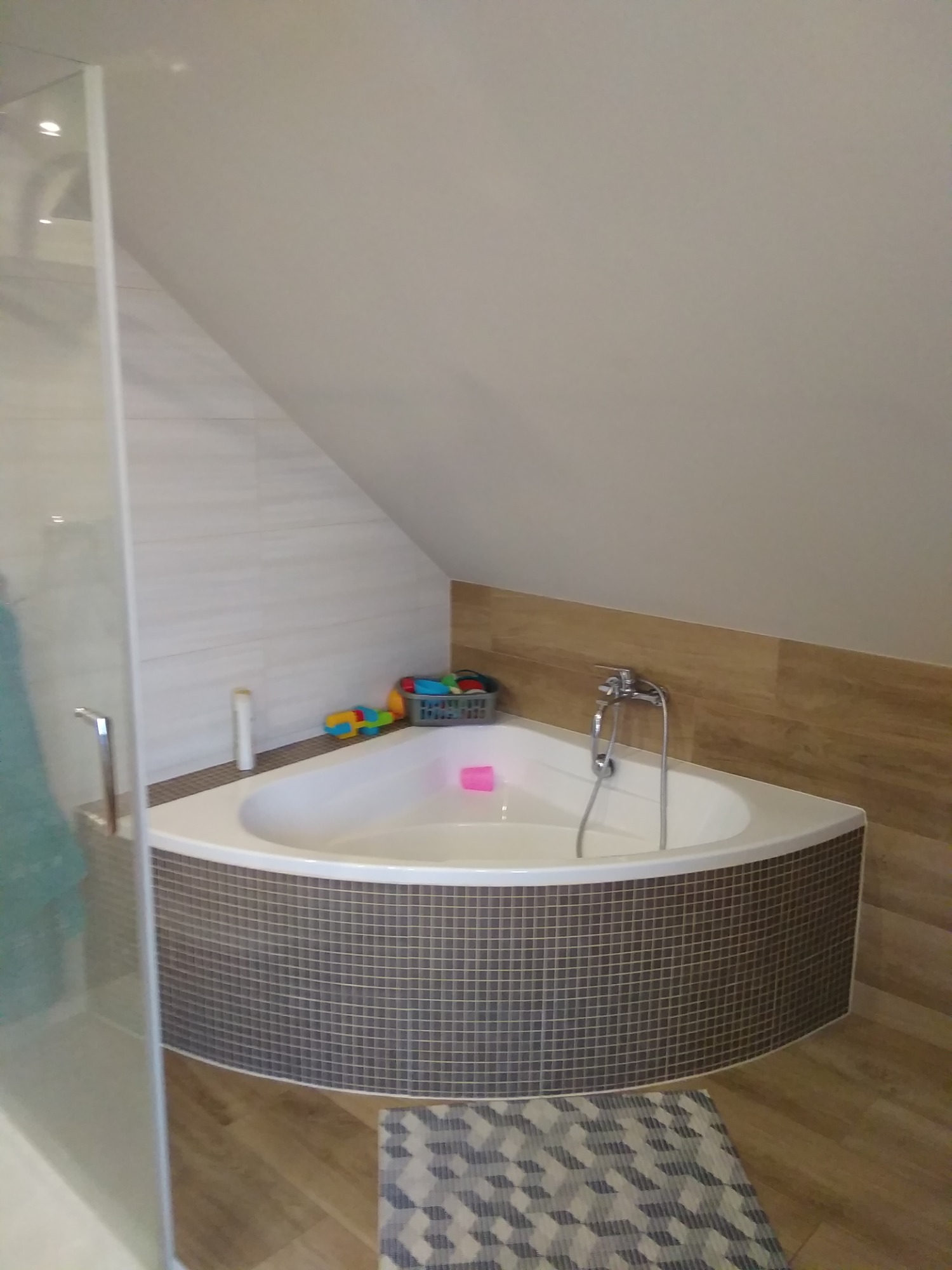 Narożna wanna obudowana mozaiką w łazience na poddaszu, z widoczną kabiną prysznicową, drewnopodobnymi płytkami i dywanikiem geometrycznym.