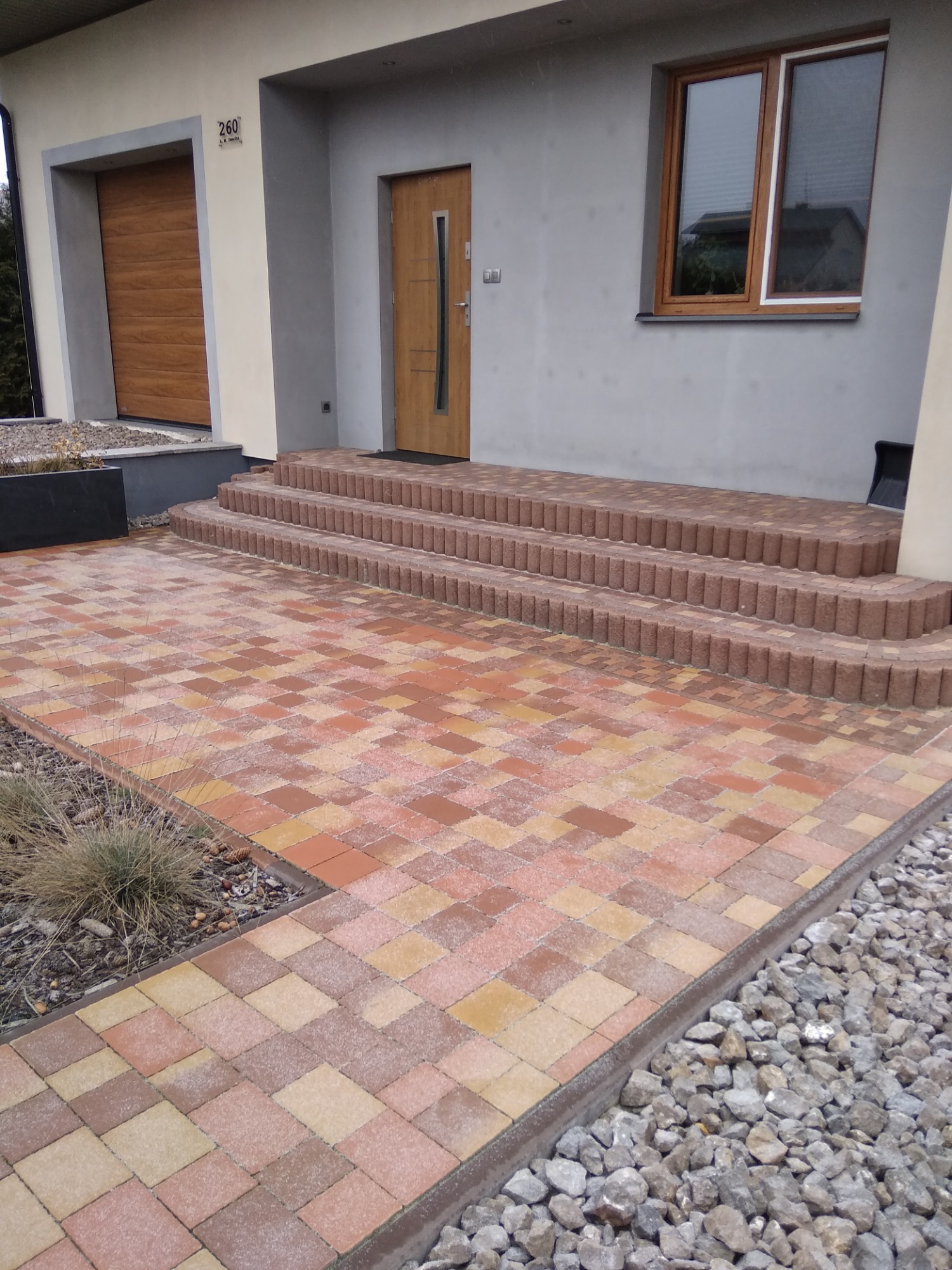 Ułożona kostka brukowa w odcieniach czerwieni i beżu prowadząca do wejścia domu w Biłgoraju, z trzema stopniami wykonanymi z cegieł. Po bokach kostki ozdobne kamienie.