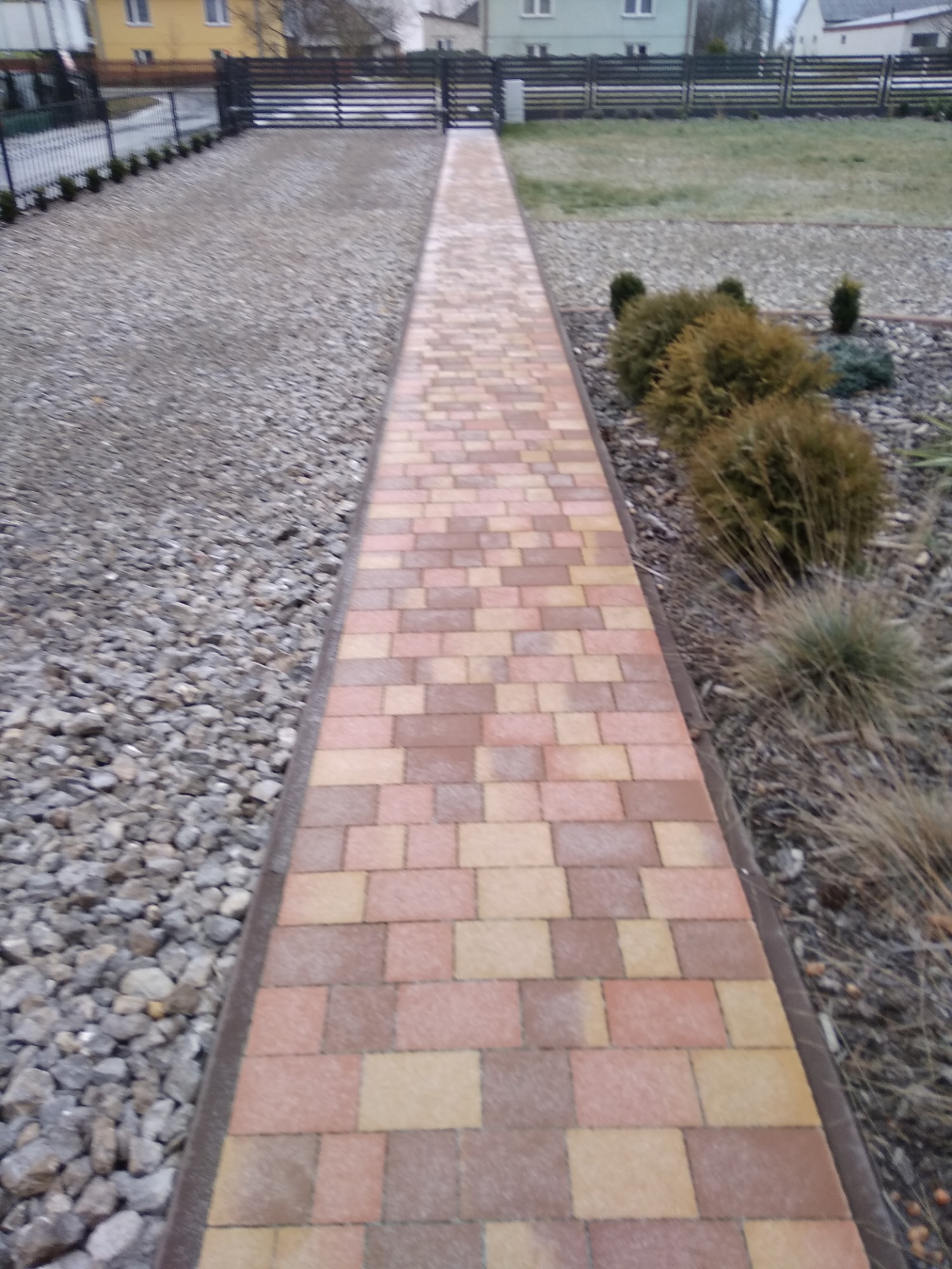 Ułożona ścieżka z kostki brukowej w odcieniach brązu i beżu prowadząca przez ogród z kamieniami i roślinami ozdobnymi, widok z perspektywy chodnika w kierunku bramy i domu w tle.