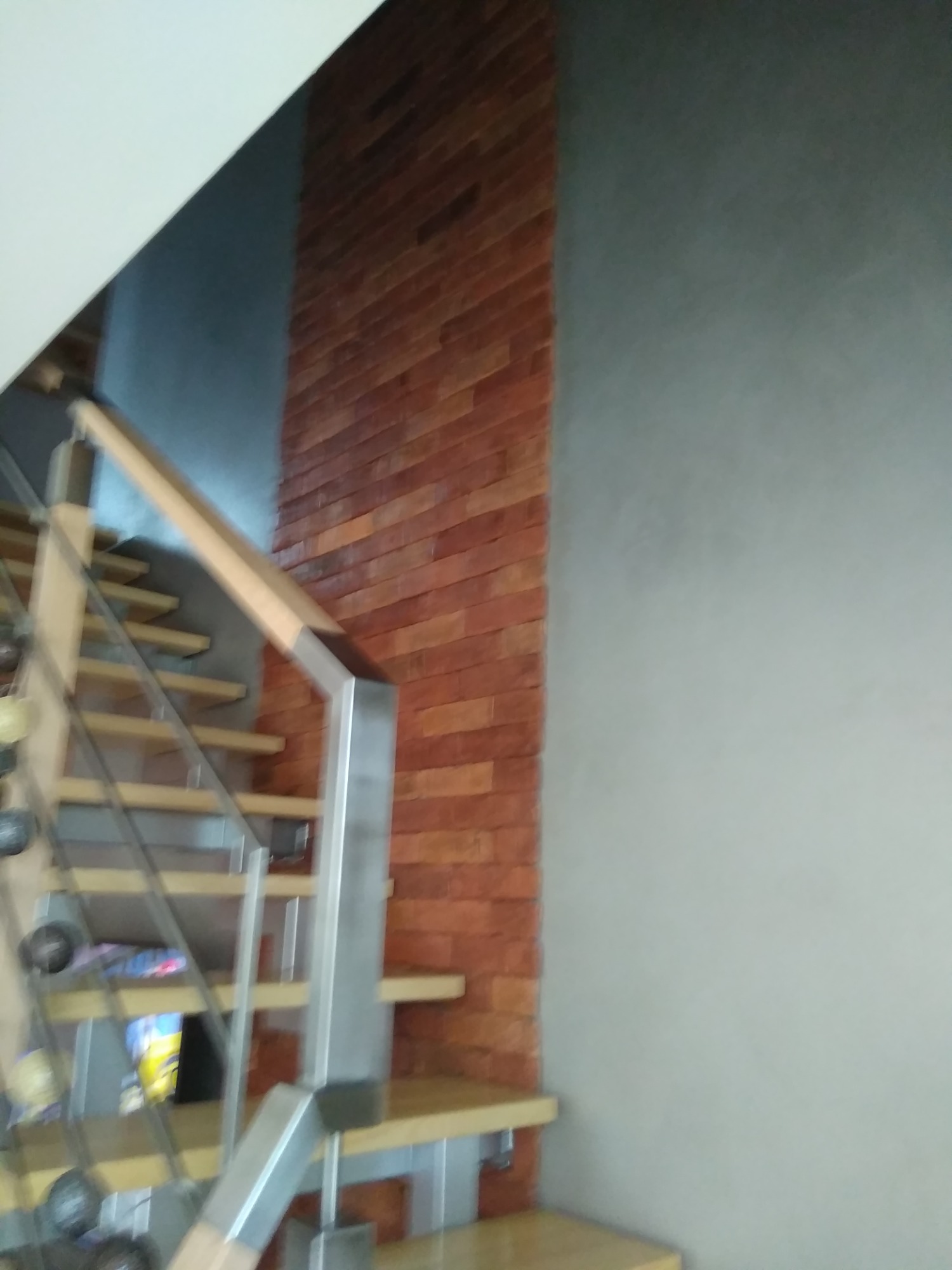 Schody z drewnianymi stopniami i metalową balustradą, przylegające do ściany wykończonej czerwoną cegłą i szarej ściany pomalowanej farbą strukturalną.