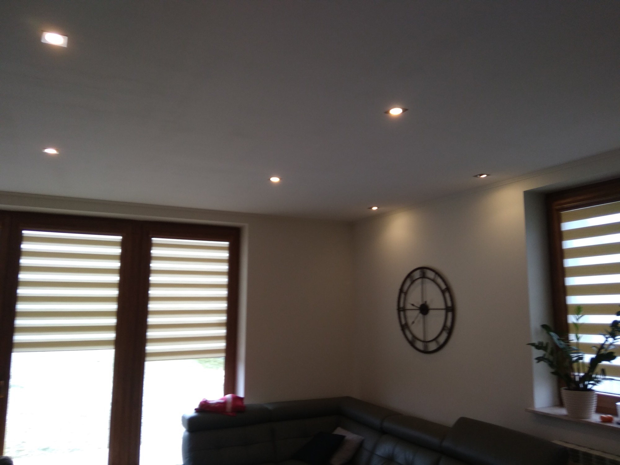 Jasne wnętrze salonu z oknami z żaluzjami dzień-noc, zegarem na ścianie i punktowym oświetleniem sufitowym.