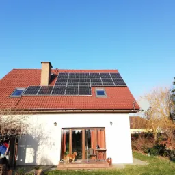 Dom jednorodzinny z czerwoną dachówką, panelami słonecznymi, oknami dachowymi, kominem i anteną satelitarną; na tarasie przed przeszklonymi drzwiami stoi stół z krzesłem i czeka pies rasy Golden...