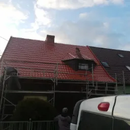 Prace na wysokości: robotnik na rusztowaniu przy czerwonym dachu z dachówki ceramicznej, obok budynek z ciemnobrązowym dachem, widoczny komin i fragment białego samochodu.