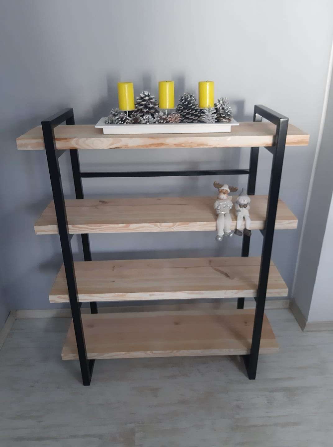 Ręcznie robiony regał z jasnego drewna i czarnych metalowych elementów, na półkach dekoracje: żółte świece, srebrne szyszki i figurki renifera i bałwanka.