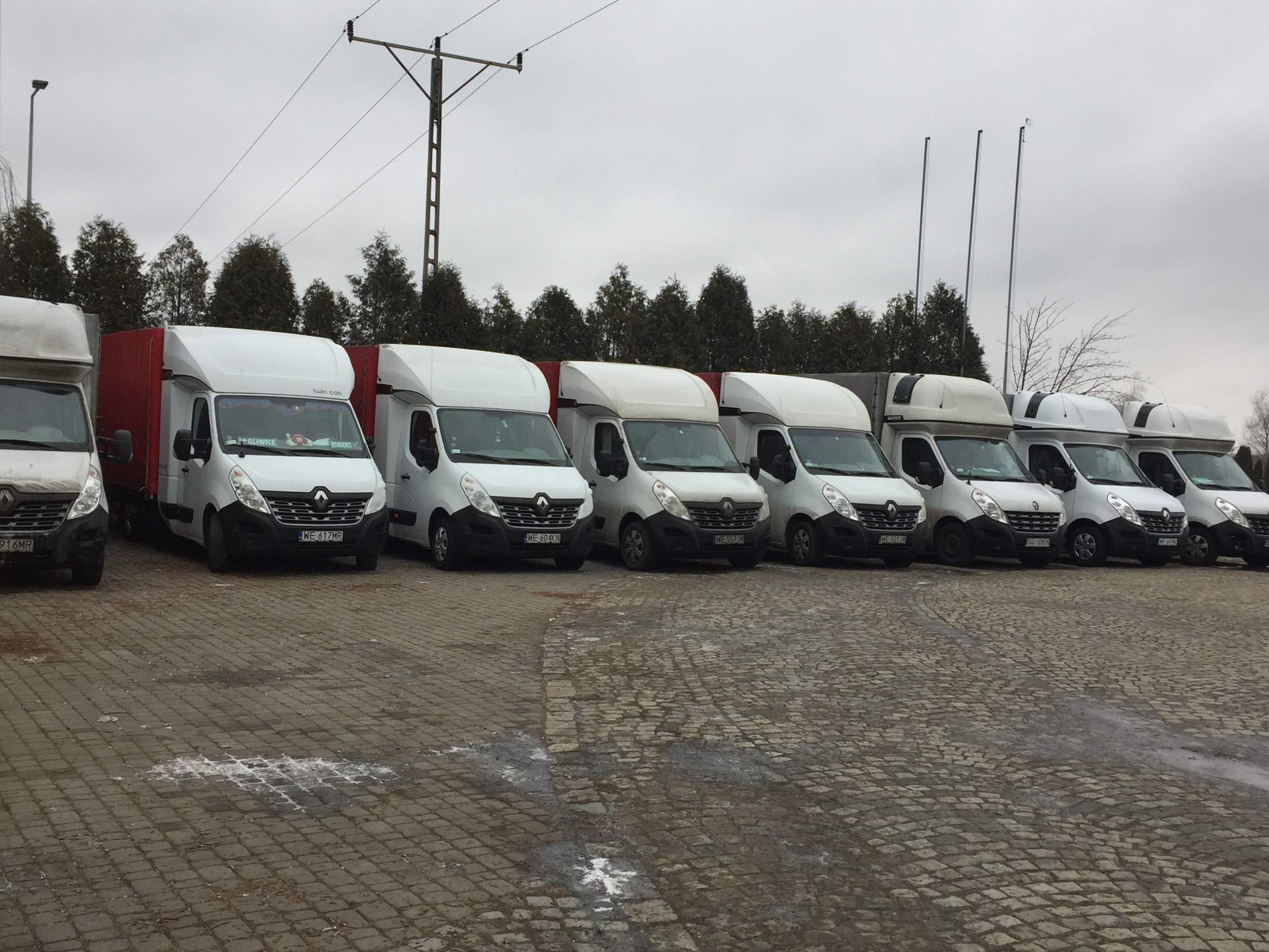 Rząd białych samochodów dostawczych Renault Master z czerwonymi i białymi zabudowami, zaparkowanych na brukowanym placu w pochmurny dzień. Na przedniej szybie pierwszego auta widoczna naklejka...