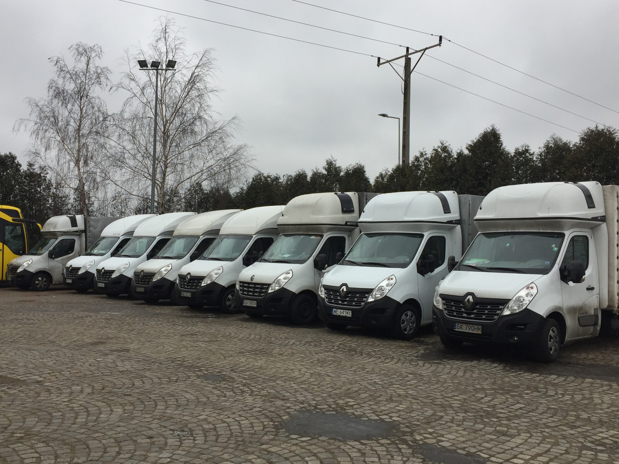 Szereg białych samochodów dostawczych Renault Master z zabudową kontenerową zaparkowanych na brukowanym placu, widok z boku, pochmurny dzień.