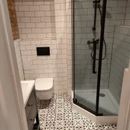 Wykończona łazienka z kabiną prysznicową w czarnej ramie, białą armaturą i wzorzystą podłogą, z widocznym fragmentem ściany z cegły w rogu.