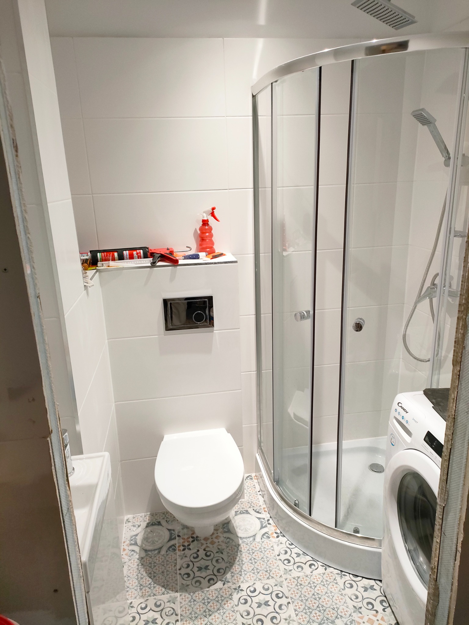 Nowoczesna łazienka w trakcie remontu: białe płytki ścienne, wzorzysta podłoga, kabina prysznicowa, toaleta podwieszana, pralka Candy i narzędzia na półce.