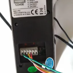 Widok z tyłu podłączonego wideodomofonu ORNO OR-VID-ME-1056KV, z widocznymi przewodami podłączonymi do złącz audio, GND, video, zasilania i zaczepów, oraz naklejką QC Passed 05 i ostrzeżeniem 'ORNO...