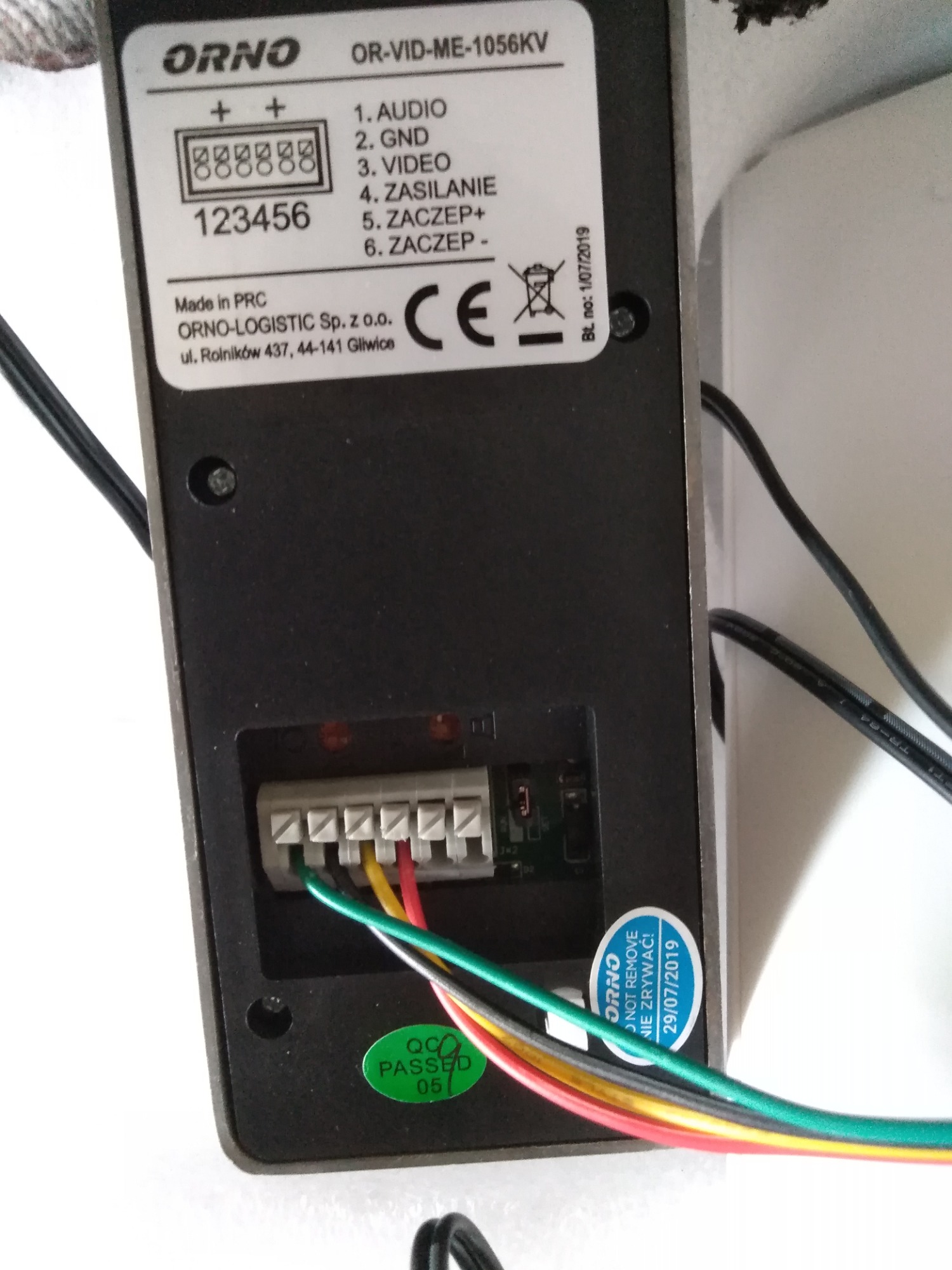 Widok z tyłu podłączonego wideodomofonu ORNO OR-VID-ME-1056KV, z widocznymi przewodami podłączonymi do złącz audio, GND, video, zasilania i zaczepów, oraz naklejką QC Passed 05 i ostrzeżeniem 'ORNO...