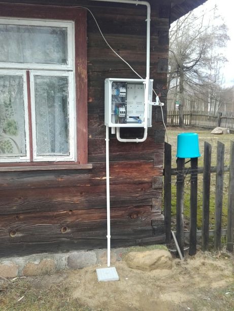 Zewnętrzna skrzynka elektryczna z bezpiecznikami, zamontowana na drewnianej ścianie domu z widocznymi przewodami i rurami PCV, na fundamencie z kostki brukowej.