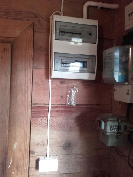 Dwupoziomowa skrzynka bezpiecznikowa zamontowana na drewnianej ścianie, obok licznik energii elektrycznej i puszka elektryczna z kablem.