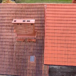 Częściowo oczyszczony dach z czerwonej dachówki, widoczny komin i okno dachowe przed i po czyszczeniu, zieleń w tle.