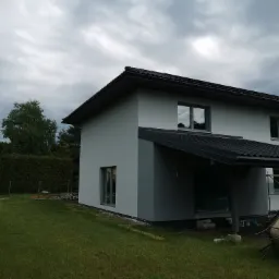 Świeżo otynkowany, dwukondygnacyjny dom jednorodzinny z ciemną dachówką, widok z boku na trawnik i pochmurne niebo.