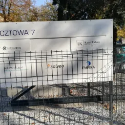 Tablica informacyjna z logotypami różnych firm, w tym kancelarii adwokackiej Karola Urbana, za metalowym ogrodzeniem, na tle jesiennego krajobrazu.