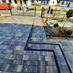 Nowo ułożona, wielobarwna kostka brukowa prowadząca od bramy wjazdowej do domu, z widocznym fragmentem buta w dolnym rogu i ekipą montażową w tle.