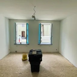 Pokój w trakcie remontu z nową podłogą z płyt OSB, otwartymi oknami zabezpieczonymi folią, skrzynką narzędziową i żółtym wiadrem na podłodze, surowa żarówka zwisa z sufitu.