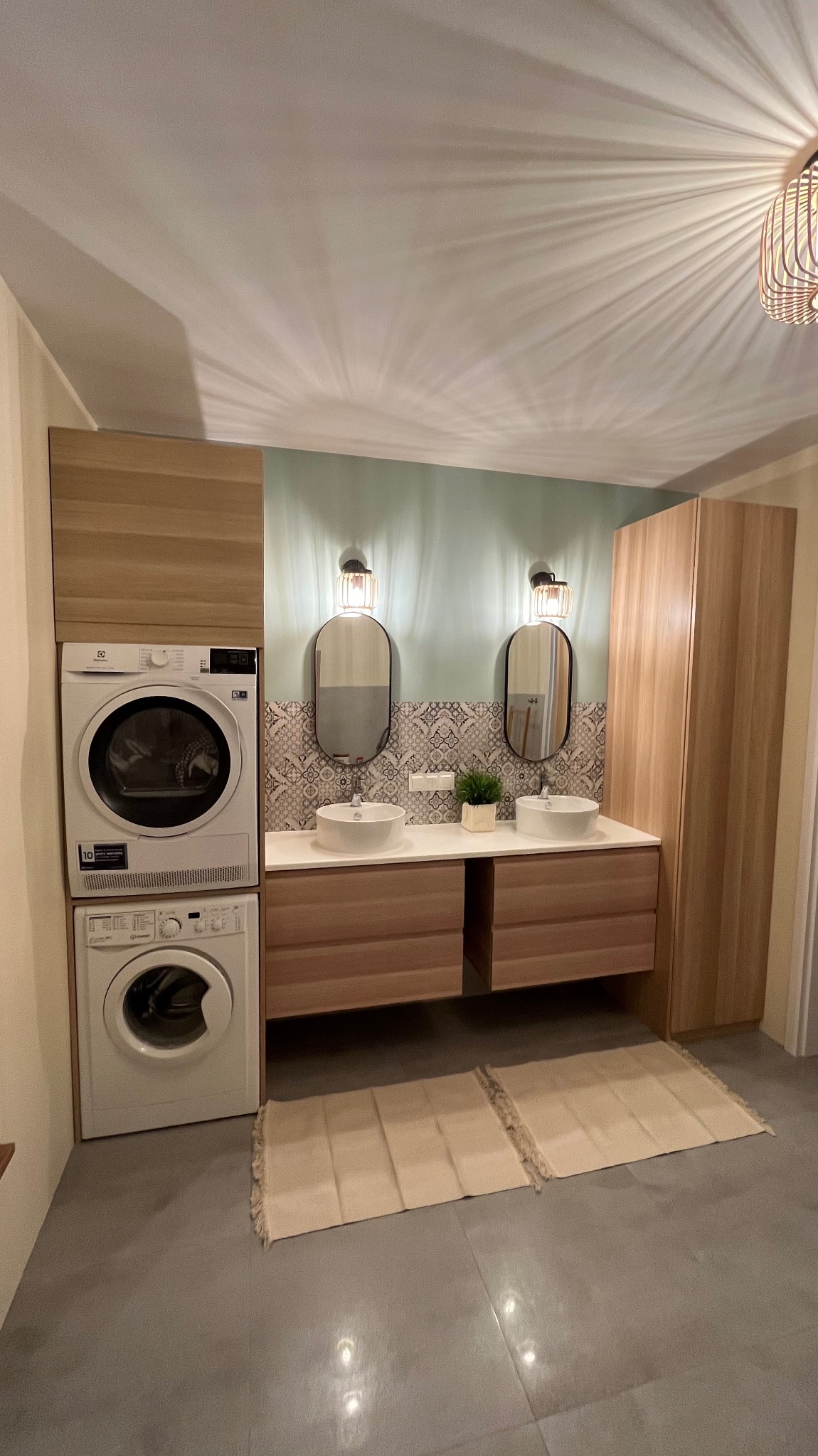 Nowoczesna łazienka z pralką i suszarką w zabudowie, dwie umywalki nablatowe z owalnymi lustrami i dekoracyjną tapetą w tle. Oświetlenie sufitowe.