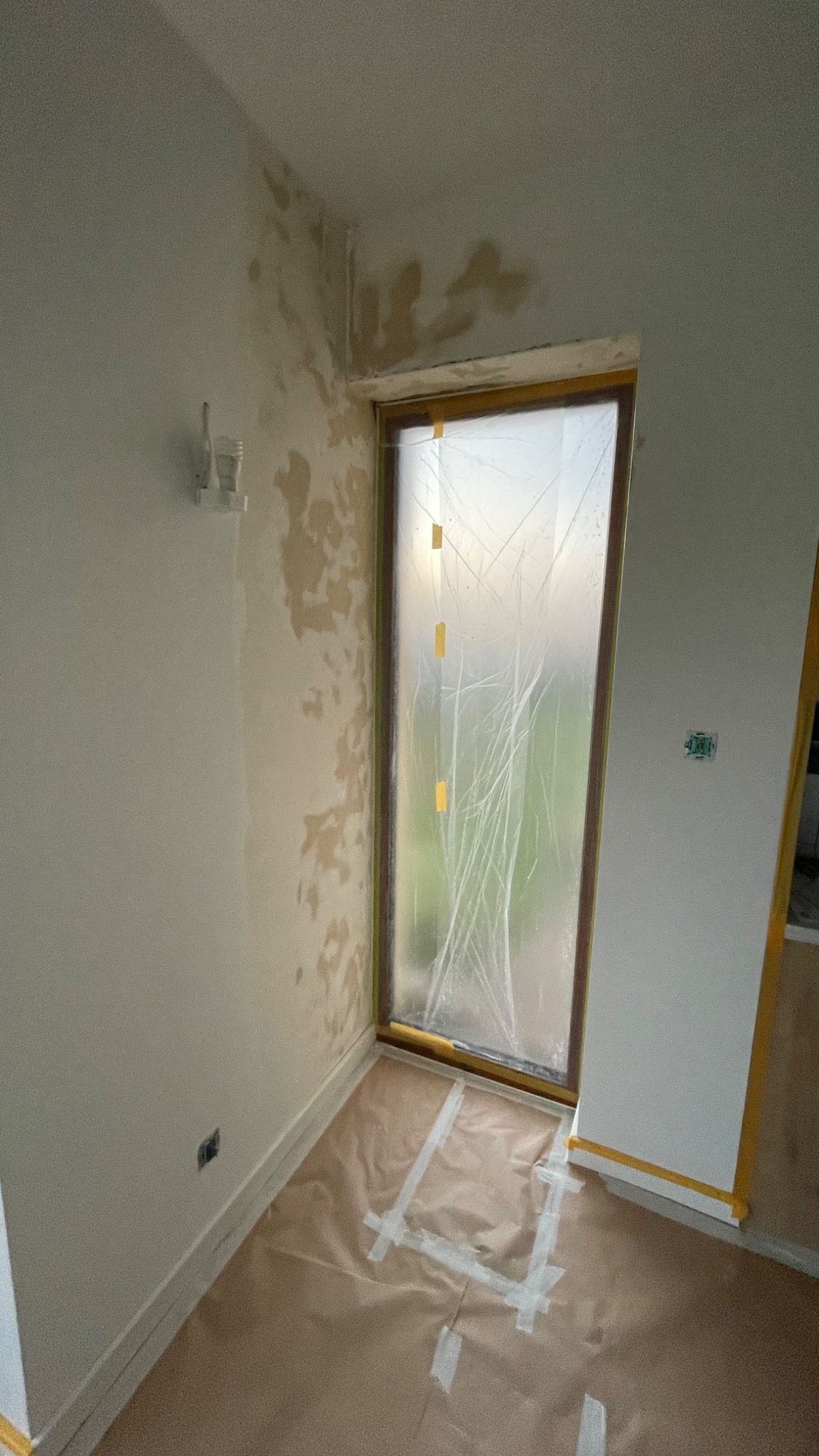 Pokój w trakcie malowania: okno zabezpieczone folią i taśmą, podłoga przykryta papierem malarskim, ściany z widocznymi śladami po wcześniejszych pracach, włącznik światła na ścianie.