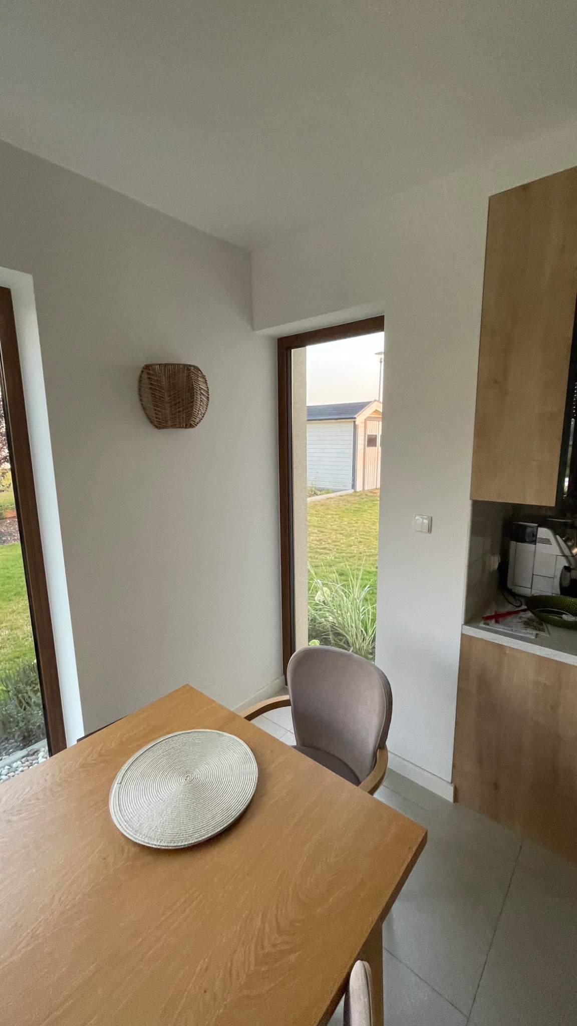 Jasne wnętrze jadalni z drewnianym stołem, plecioną podkładką i krzesłem na tle okna z widokiem na ogród i budynek gospodarczy.