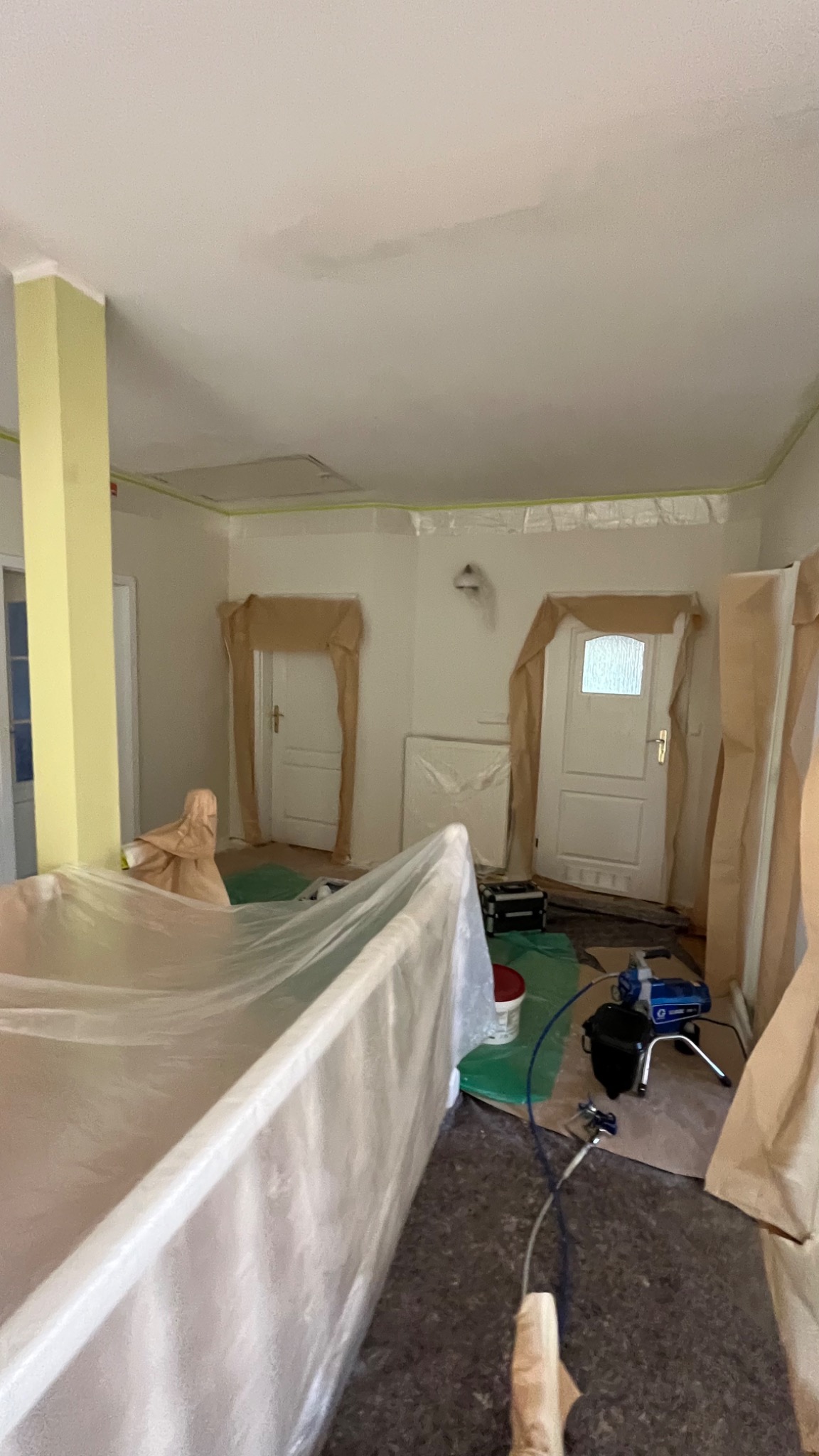 Pokój przygotowany do malowania natryskowego: folia ochronna na meblach i drzwiach, taśma malarska przy suficie, agregat malarski na podłodze.