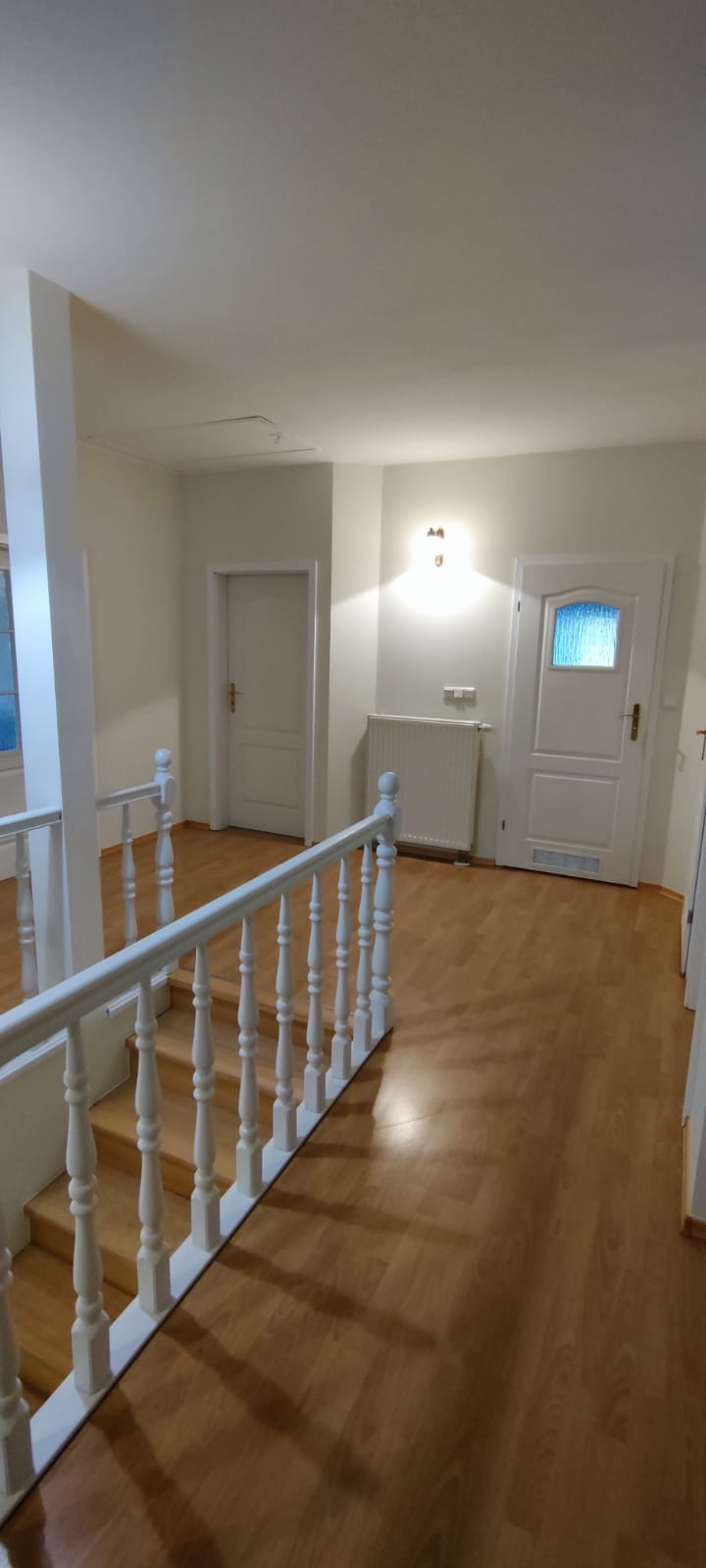 Jasne wnętrze domu z drewnianą podłogą, białymi ścianami, schodami z białą balustradą i dwoma białymi drzwiami, z których jedne mają szybę z niebieskim szkłem.