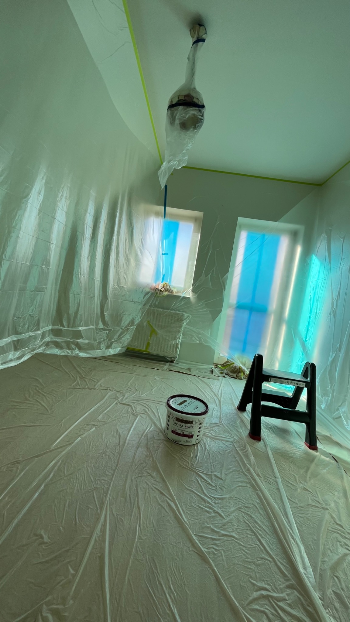 Pokój przygotowany do malowania: folia ochronna na podłodze i oknach, taśma malarska na suficie, wiadro z farbą Ultra Matt i mała drabinka.