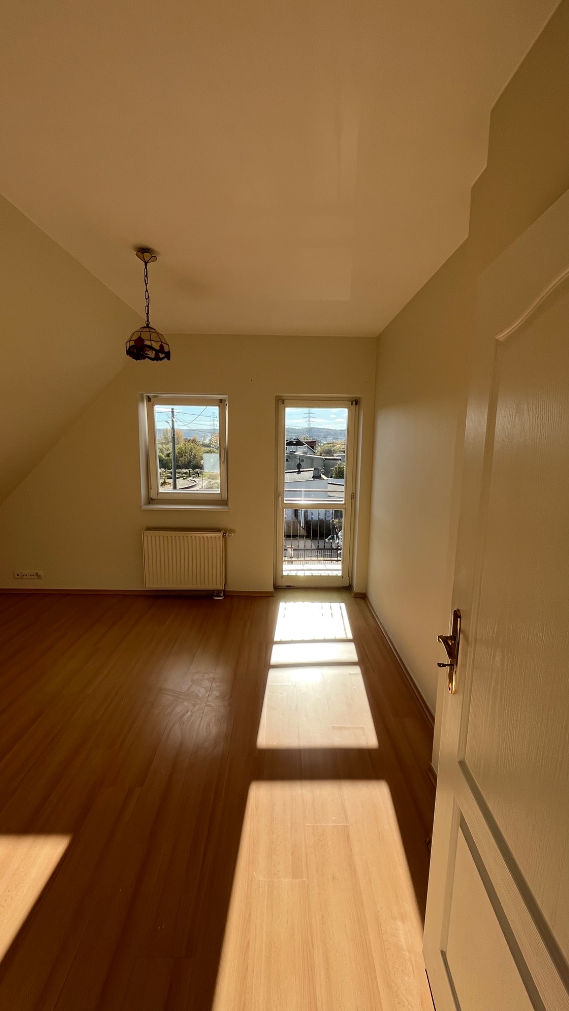 Puste, jasne poddasze z oknem i drzwiami balkonowymi, parkiet na podłodze, światło słoneczne tworzące długie cienie, widoczny grzejnik pod oknem, lampa wisząca z kolorowym abażurem.